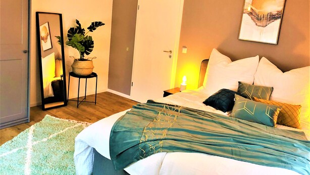 A bedroom with a bed, bedside table, lamp, potted plant, and a painting on the wall.