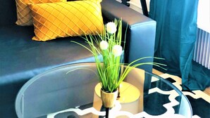 A modern living room with a dark sofa, yellow cushions, a glass coffee table with a potted plant, a floor lamp, and a blue curtain.