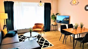 A modern living room with a sofa, coffee table, dining table, and chairs. There is a TV on the wall and a plant on a side table.