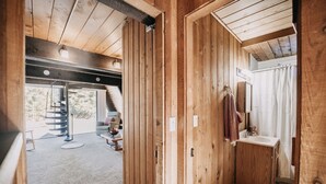 A wooden interior with a staircase leading to a balcony, a bathroom with a sink and towel rack, and a seating area with a table and chairs.