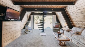 A cozy, wooden interior with a television, a rocking chair, a sofa, and a spiral staircase leading to an outdoor area.