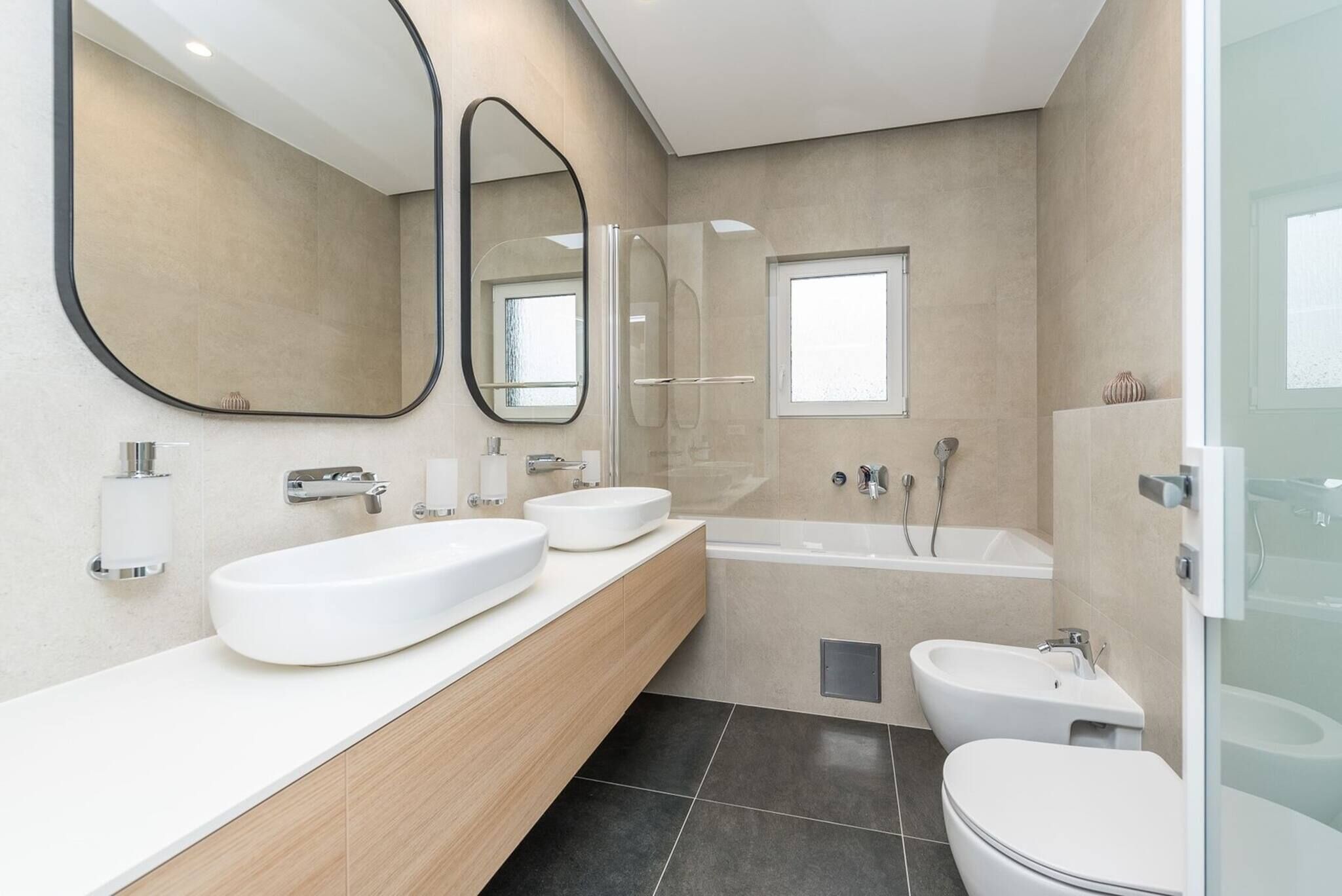A modern bathroom with a large mirror, two sinks, a bathtub, and a toilet.