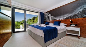 Quarto moderno com uma cama grande, cabeceira de madeira, mesa de cabeceira e vista para o verde através de uma porta de vidro deslizante.