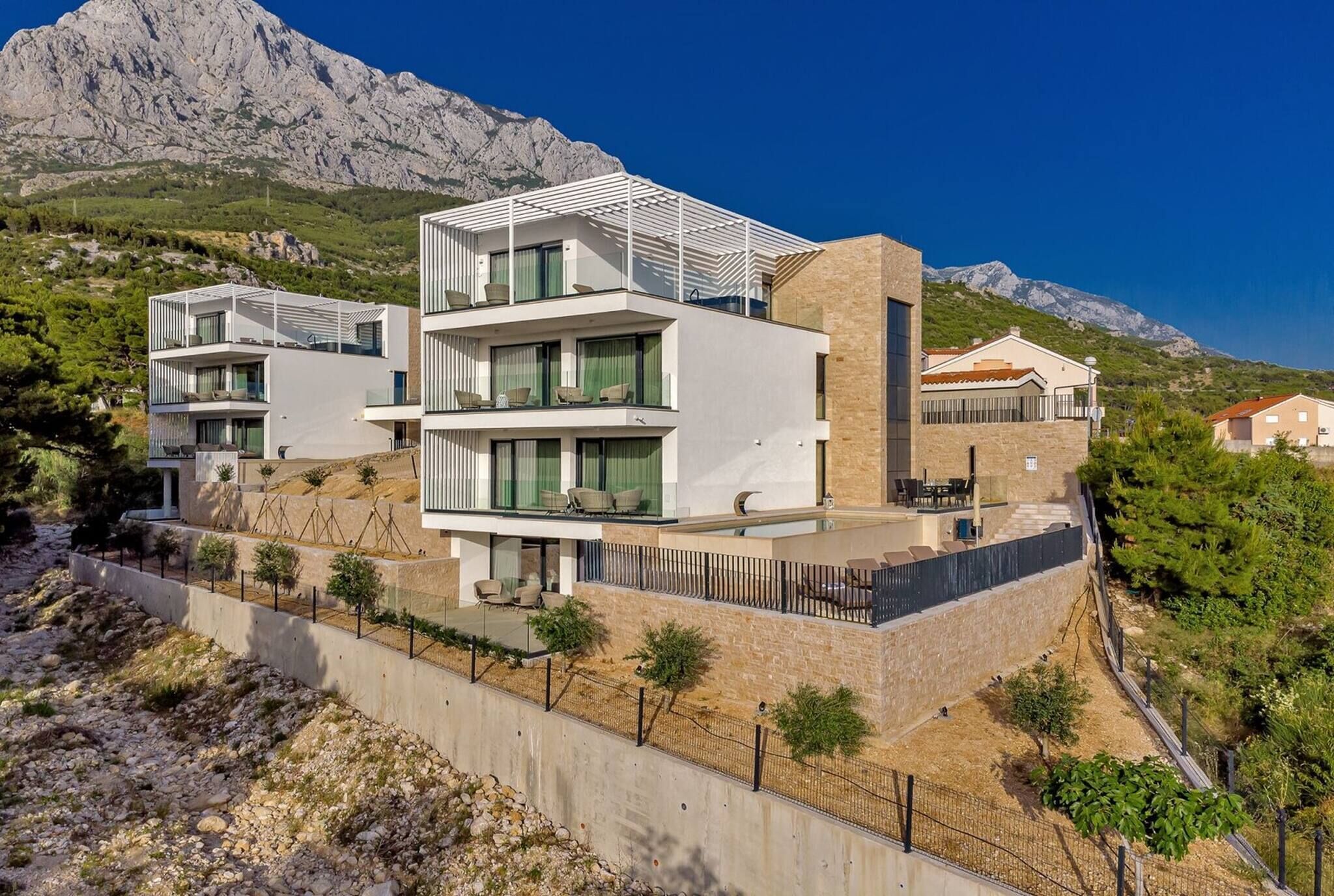 A modern multi-story building with balconies, a stone retaining wall, and a mountainous backdrop.