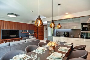 Un comedor moderno con una mesa larga dispuesta para cuatro, iluminación colgante, un televisor al fondo y una cocina con microondas y cafetera.