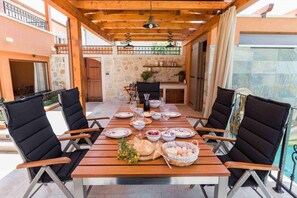 Un área de comedor al aire libre techada, con una mesa larga, sillas y un muro de piedra de fondo.