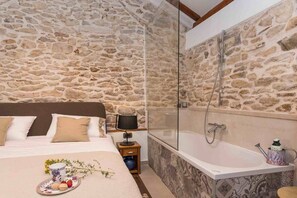 Un dormitorio con pared de piedra, una cama grande con almohadas, una mesita de noche con lámpara, y un baño con bañera y ducha.