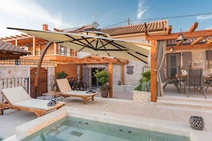 Un espace piscine aménagé avec des chaises longues, une pergola et un bâtiment en pierre surmonté d’un toit en tuiles.
