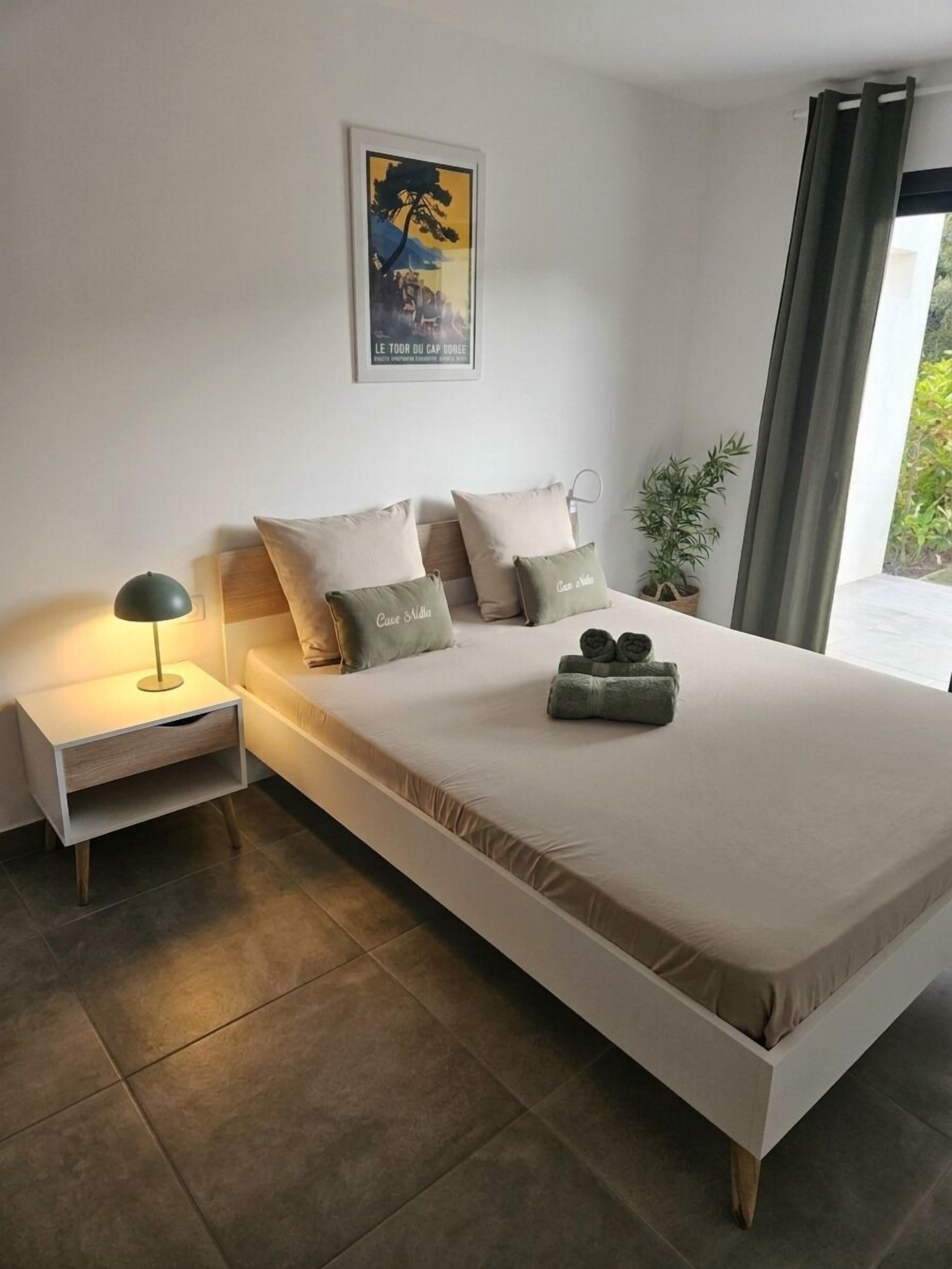 A modern bedroom with a bed, bedside tables, a lamp, a potted plant, and a painting on the wall.