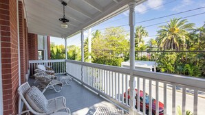 A covered porch with wicker furniture, a white railing, and a view of palm trees and parked cars.
