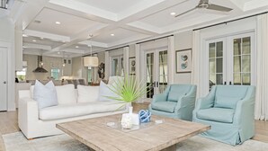 Amplia sala de estar con un sofá blanco, dos sillones azul claro, una mesa de centro de madera y un ventanal con contraventanas.