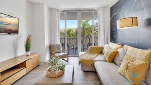 A modern living room with a sofa, a chair, a TV, a plant, and a balcony with a view. - Modern Resort:   Awesome Pool + Gym|Near Beach (Hallandale Beach)