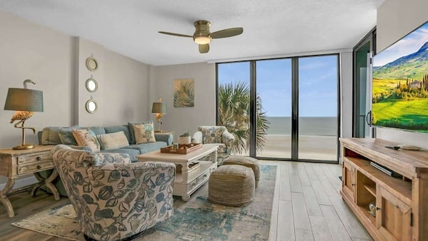 A spacious living room with a large sliding glass door leading to a balcony, a ceiling fan, and a flat-screen TV. - Oceanfront, Pool, Beach Gear, Tennis, Smart TVs (St. Augustine)