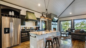 A modern kitchen with a large island, stainless steel appliances, and a stone fireplace. - Treetop Views for Miles! New, Romantic Couples Cabin (Broken Bow)