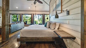 A spacious bedroom with a large bed, wooden beams, a ceiling fan, and a view of greenery outside. - Treetop Views for Miles! New, Romantic Couples Cabin (Broken Bow)