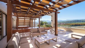 A sunlit outdoor seating area with a wooden pergola, cushioned sofas, and a view of the landscape.