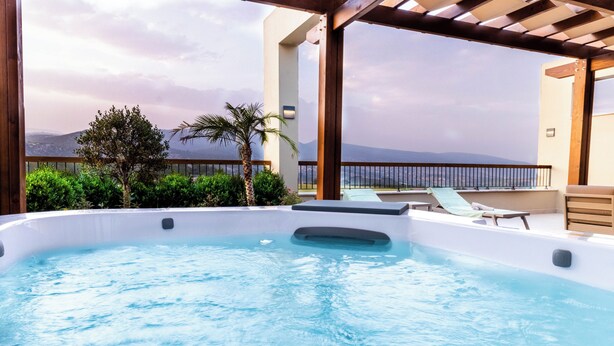 A hot tub with a view of the sea and mountains, surrounded by greenery and a wooden deck.