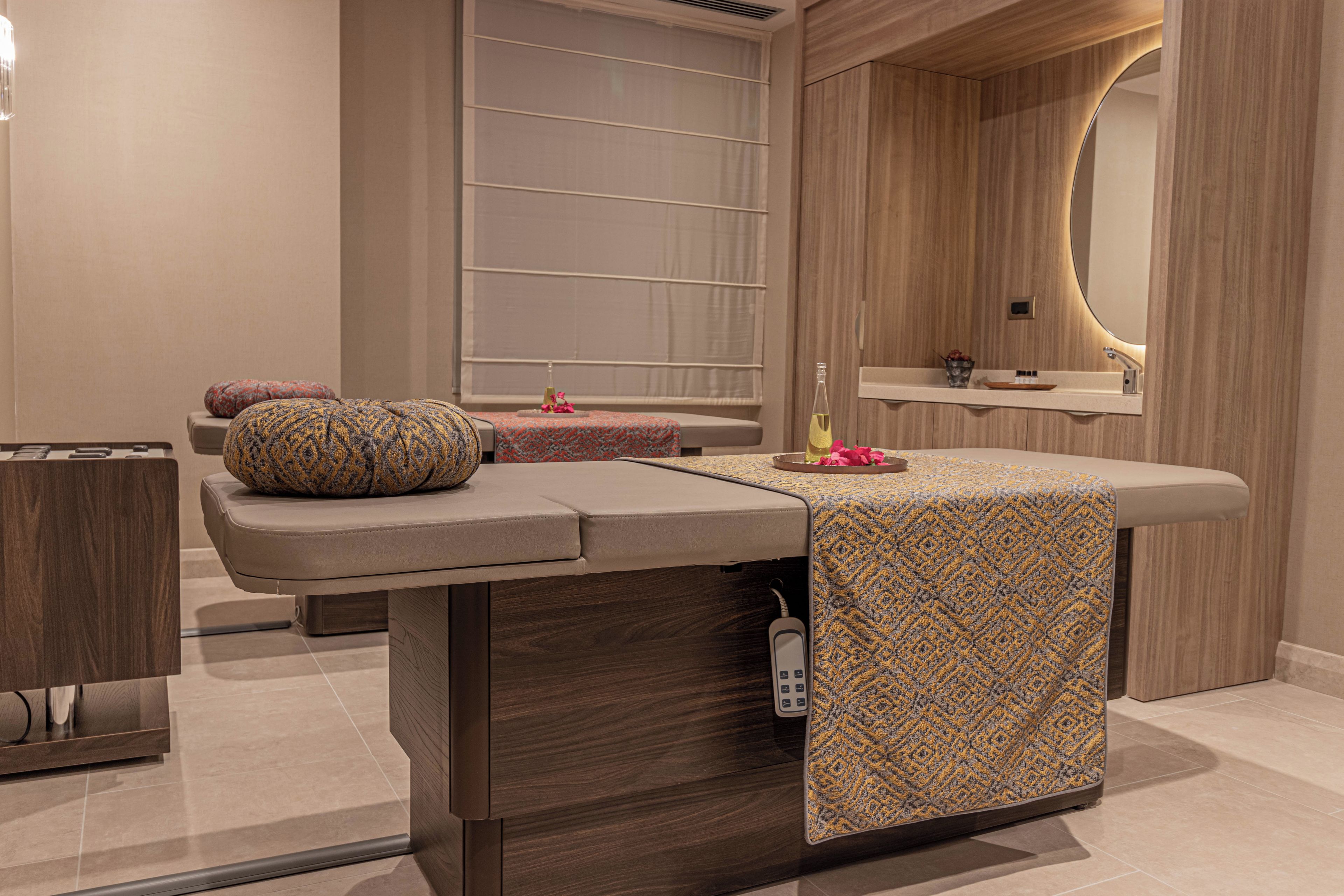 Ett modernt behandlingsrum för spa med en massagebänk, ett litet bord med en flaska och blommor samt en spegel med en träram.