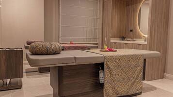 Ett modernt behandlingsrum för spa med en massagebänk, ett litet bord med en flaska och blommor samt en spegel med en träram.
