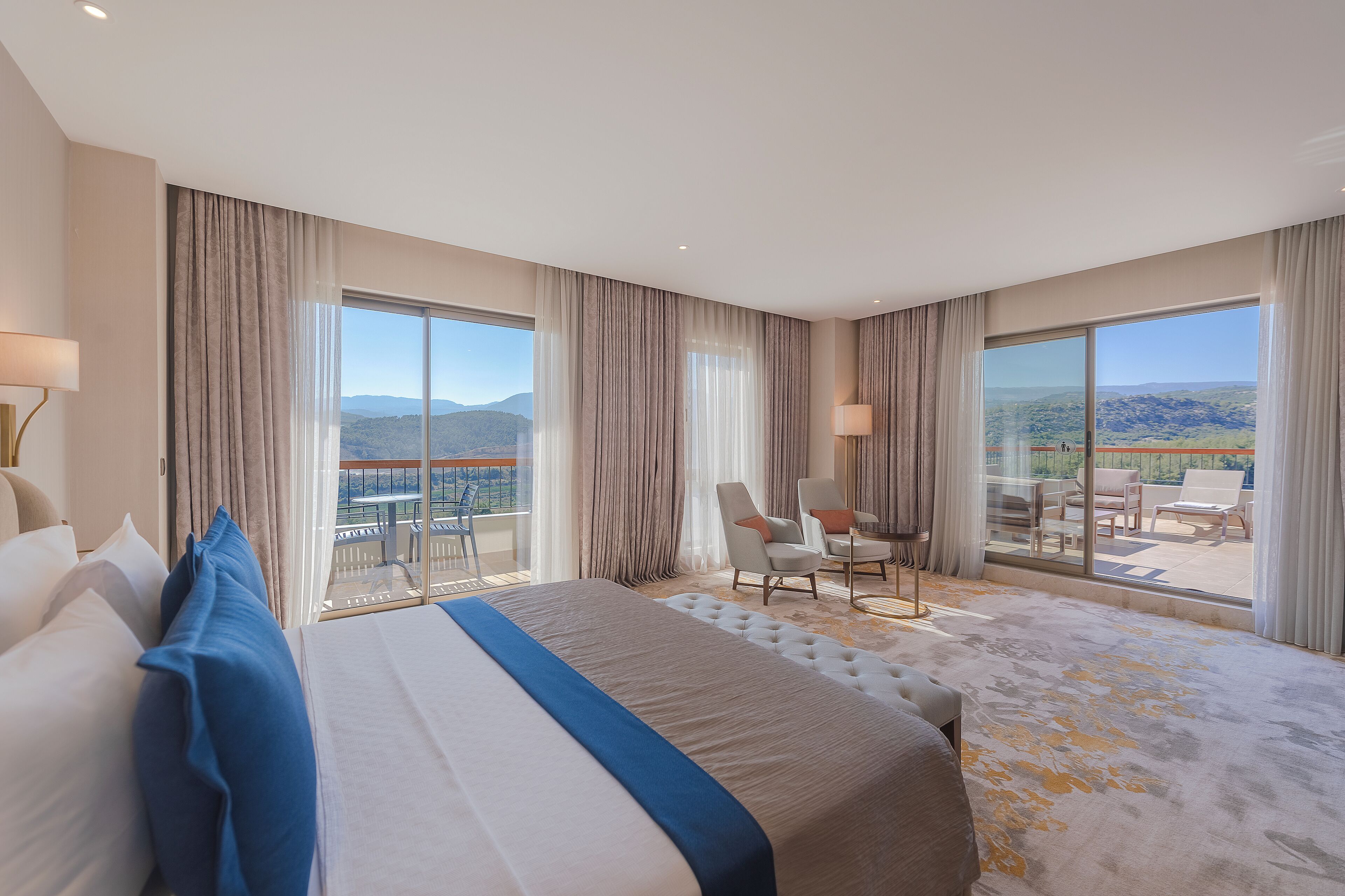 A hotel room with a large bed, two chairs, a balcony with a table, and a view of the mountains.