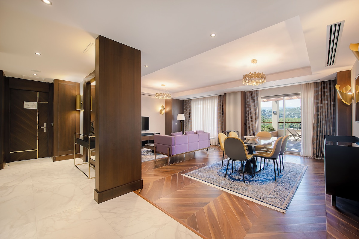 Une chambre d’hôtel moderne avec un plancher en bois, un canapé violet, un coin repas avec une table et des chaises, et une grande fenêtre avec des rideaux.