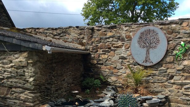Un mur de pierre orné d’un motif d’arbre, une statue de paon et un pot bleu contenant des plantes.