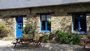 Un bâtiment en pierre avec des portes et des fenêtres bleues, une terrasse en gravier avec des tables de pique-nique et un jardin fleuri.