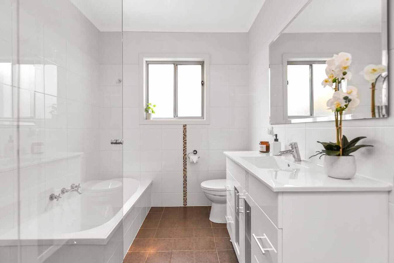 A modern bathroom with a bathtub, toilet, and sink. There is a window above the toilet and a mirror above the sink.