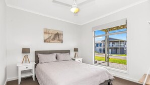 Un dormitorio con una cama, dos mesitas de noche, un ventilador de techo, una ventana con vista a un edificio moderno y un cuadro en la pared.