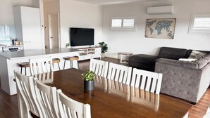 Amplia sala de estar con una mesa de comedor de madera rodeada de sillas blancas, un sofá gris y un televisor de pantalla plana.