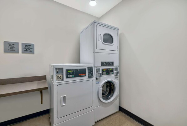 Un cuarto de lavandería con lavadoras y secadoras apiladas, una recepción y carteles que indican los tiempos de ciclo y la disponibilidad de productos.