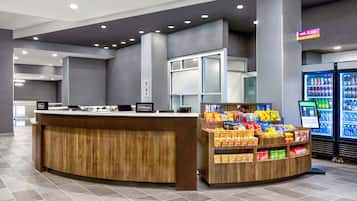 Un espace d’accueil d’hôtel avec un comptoir en bois, un distributeur de boissons en libre-service et un écran numérique affichant des informations.