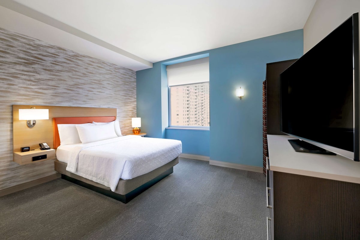 Ein Hotelzimmer mit Bett, Nachttisch, Lampe, Fernseher und einem Fenster mit Blick auf Gebäude.