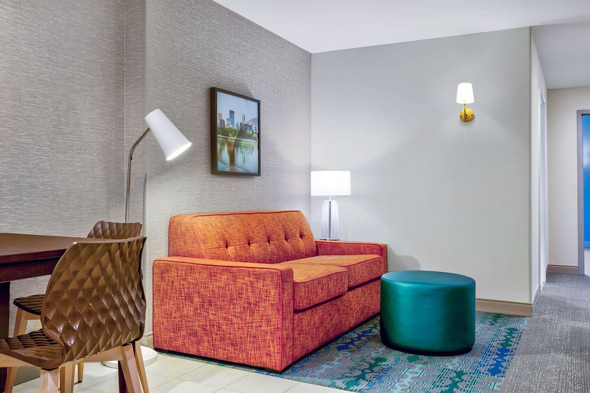 Ein modernes Hotelzimmer mit einem roten Sofa, einem petrolfarbenen Hocker, einem Esstisch mit Stühlen und einem Stadtbild an der Wand.