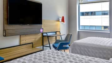 A hotel room with a bed, a desk, a chair, a TV, and a window with a view of buildings.