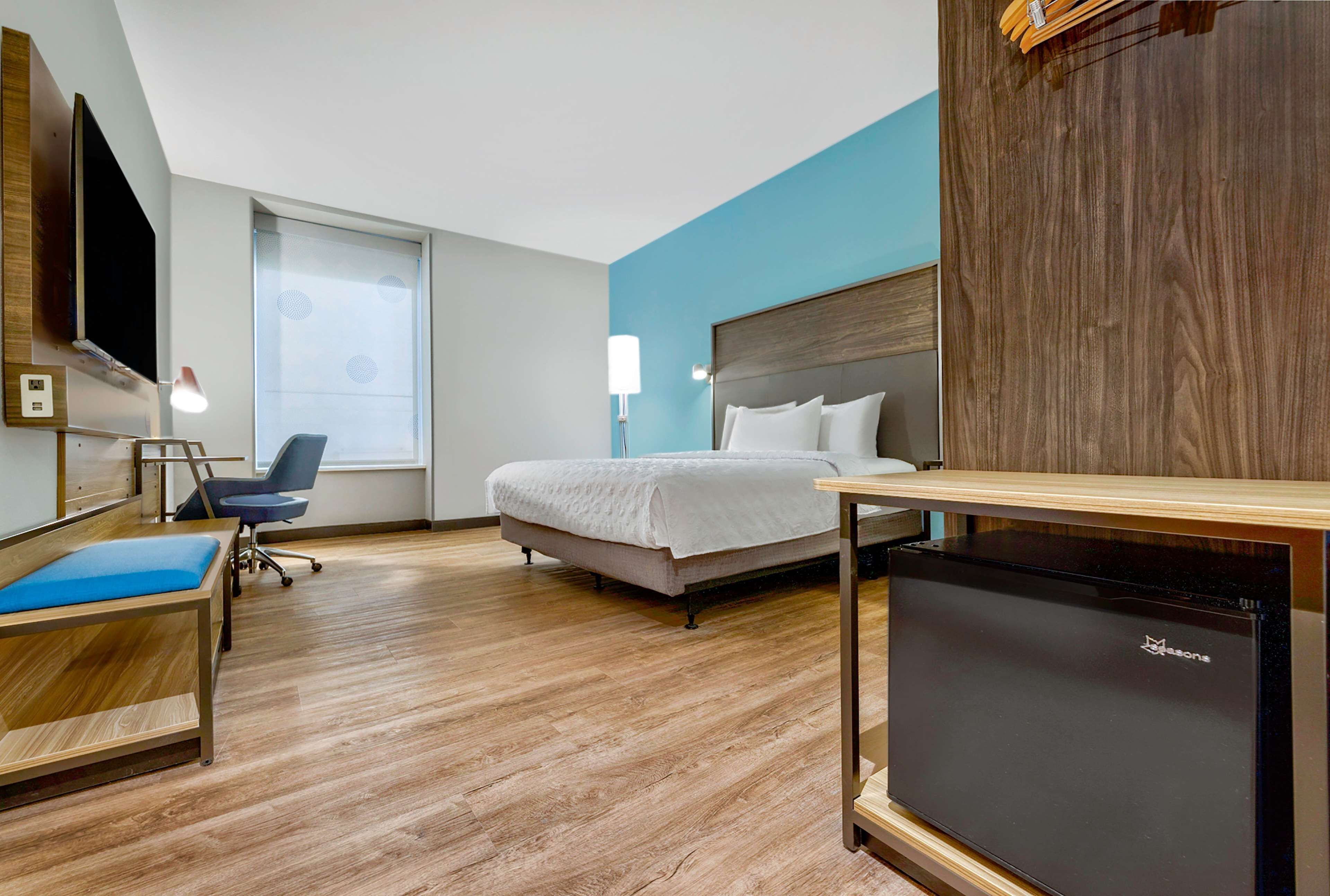 A hotel room with a bed, a desk, a chair, a TV, and a refrigerator.