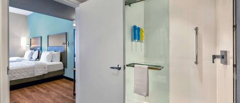 A modern hotel room with a wooden floor, a bed with white bedding, a glass shower enclosure, and a glass door.