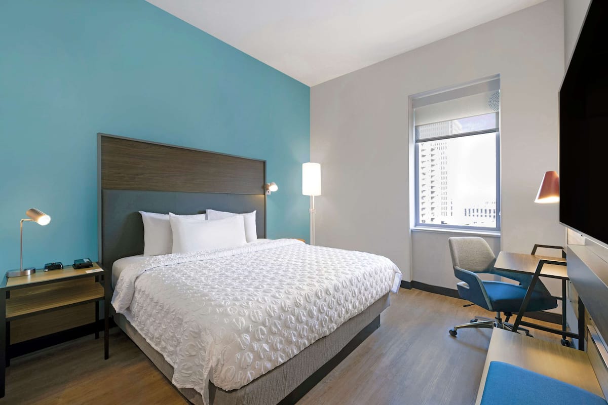 Habitación de hotel con cama, mesita de noche, lámpara, silla, escritorio y televisor.