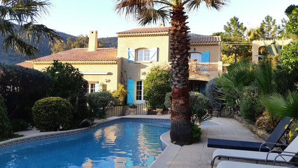 Un espace piscine avec une piscine carrelée, des chaises longues et une maison avec un toit en tuiles et des volets bleus.