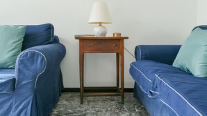 Una stanza con due divani blu, uno con un cuscino color petrolio, e un comodino in legno con una lampada bianca e un libro.