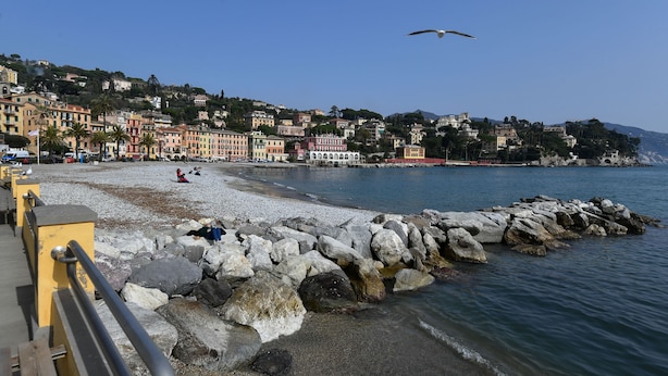 Una località costiera con una spiaggia rocciosa, una fila di edifici colorati e un cielo azzurro limpido.