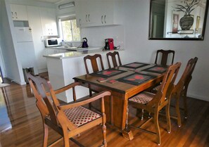 A dining area with a wooden table and chairs, a kitchen in the background, and a vase on the wall. - Eagles Nest Cottage where everything is better with an ocean view and breeze (Crowdy Head)