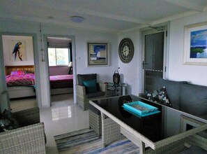 A room with a bed, a sofa, a coffee table, and a dining table. There is a window with a view of trees and a door leading to another room. - Eagles Nest Cottage where everything is better with an ocean view and breeze (Crowdy Head)