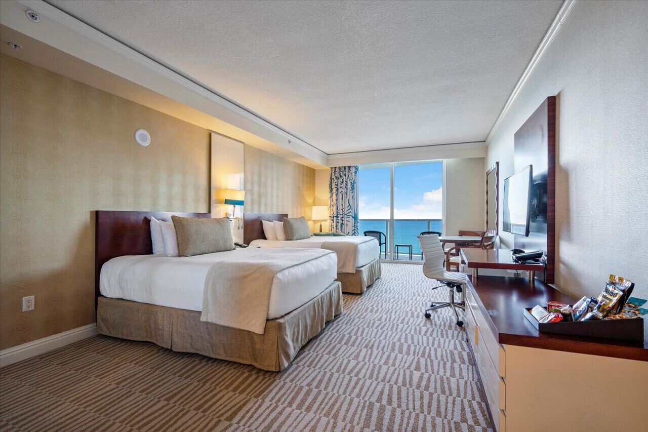 Habitación de hotel con dos camas, un escritorio, una silla y un ventanal con vista al mar.