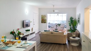 A modern living room with a sofa, dining table, and a TV. There are plants and a window with natural light. - Beauty Bavaro Playa Matilde (Punta Cana)