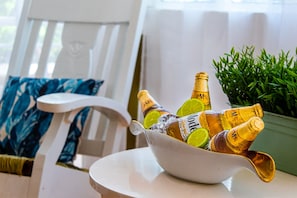 A white chair with a blue floral cushion, a white table with a bowl of champagne bottles and lime slices, and a green plant in the background.