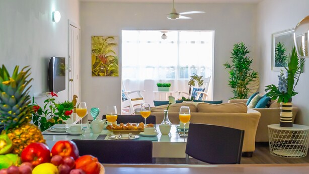 A bright living room with a sofa, dining table, and chairs. There are potted plants and a TV mounted on the wall. - Beauty Bavaro Playa Matilde (Punta Cana)