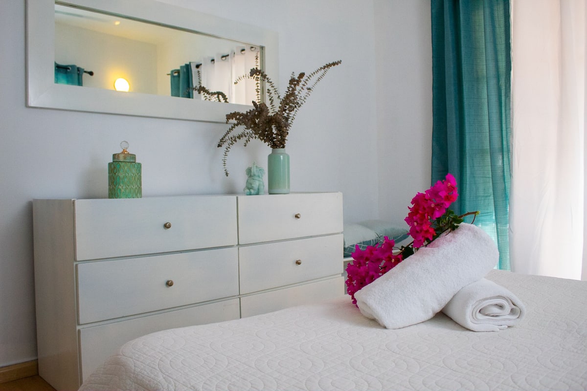 A neatly made bed with a white quilt, a white dresser with drawers, a vase with dried plants, and a pink flower arrangement.