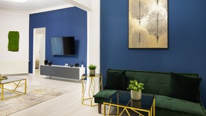A modern living room with a blue accent wall, a green sofa, a gold-framed coffee table, and a flat-screen TV mounted on the wall. - Metropole Apartments Old Center (Bucharest)