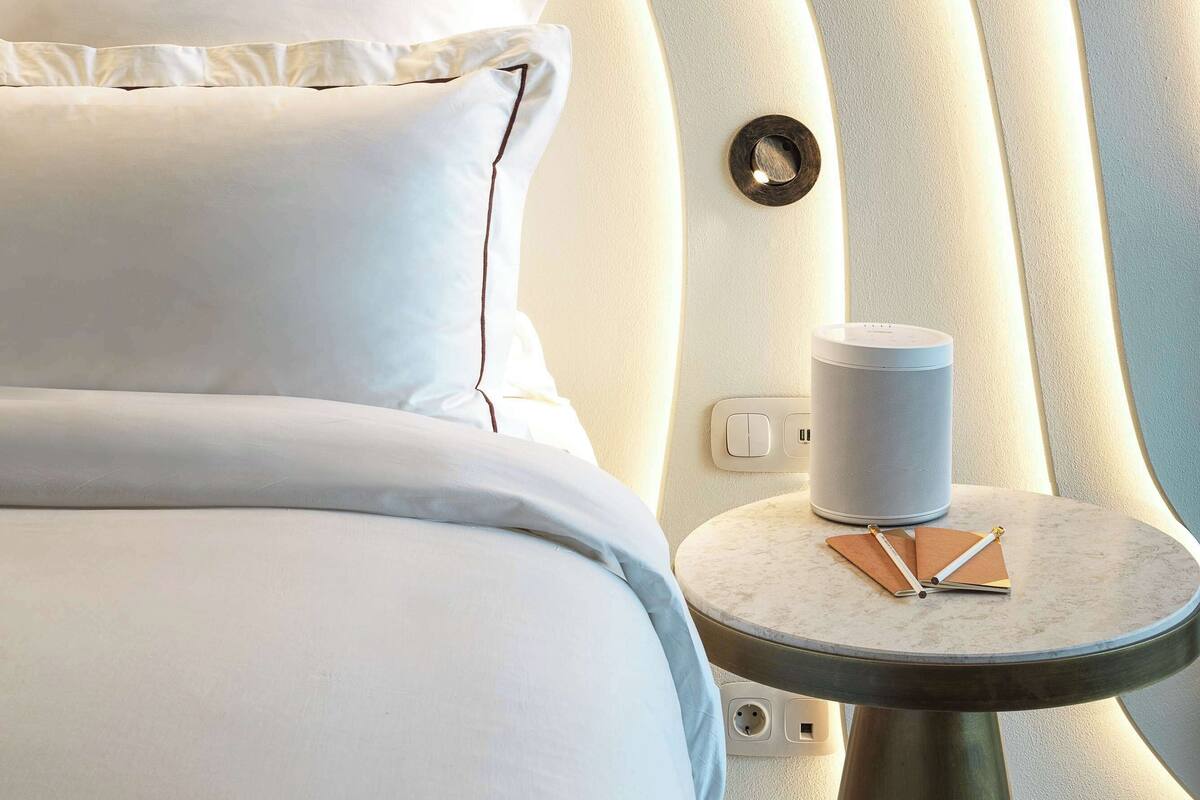 Un lit moderne avec du linge de lit blanc et une table de chevet ronde, sur laquelle se trouvent une lampe et un livre.